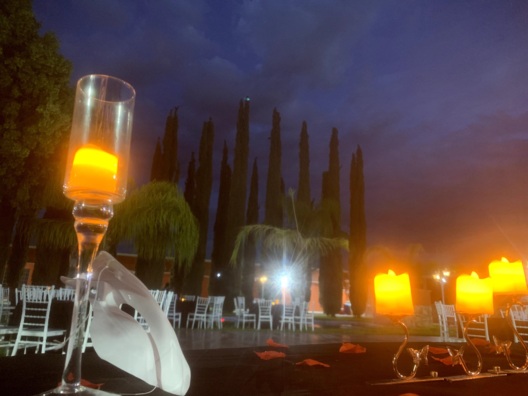 Vela en copa de champán sobre una mesa con mantel negro, rodeada de velas y sillas blancas en el jardín de Quinta Paris.
