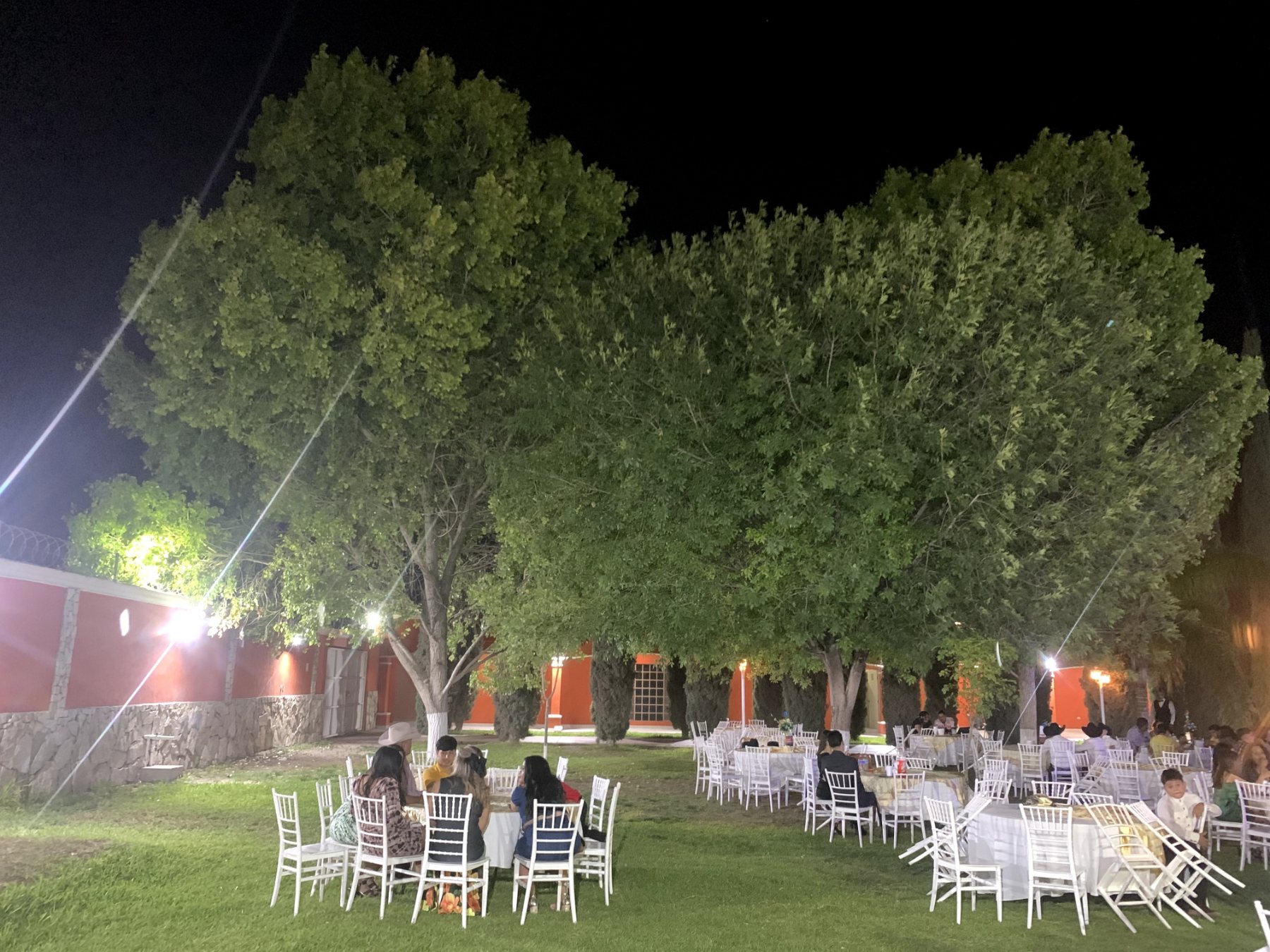 Jardín iluminado de noche con mesas y sillas blancas, y personas sentadas Quinta Paris Lerdo